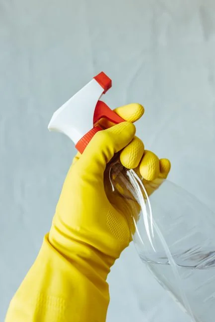 Homme anonyme portant des gants en latex jaunes et un t-shirt sombre, essuyant un miroir dans une salle de bain avec une serviette en papier